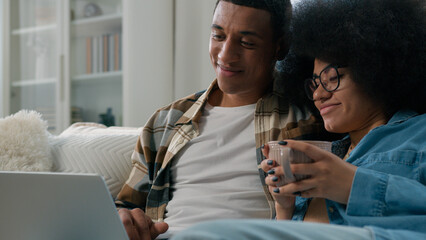 African American couple at home couch looking at laptop talking watching film movie discuss...