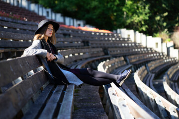 A lonely girl in a black hat sits in on a summer theater bench.