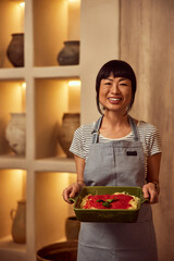 Portrait of a smiling Asian woman who made dinner for her friends, wearing an apron and holding a dish. - 756408601
