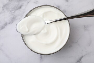 Delicious natural yogurt in bowl and spoon on white marble table, top view