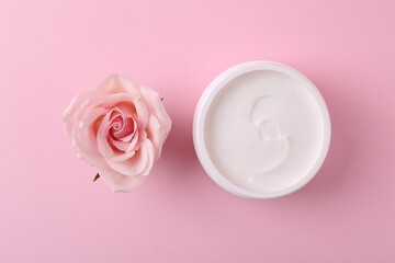 Moisturizing cream in open jar and rose flower on pink background, flat lay. Body care product