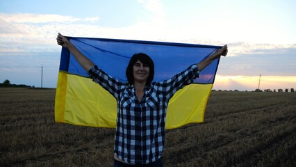Happy woman looks into camera standing on wheat field with a lifted blue-yellow banner at sunset. Ukrainian smiling lady posing with a raised over head flag of Ukraine on barley meadow at sunrise. - 756420271