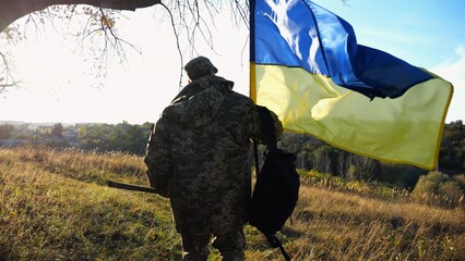 Young male military in uniform walks with waving flag of Ukraine at countryside. Soldier of ukrainian army going with blue-yellow banner outdoor. Concept of victory against russian aggression. - 756420665