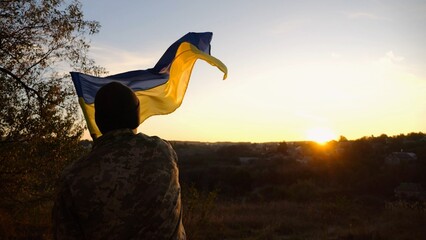 Young man in military uniform waving flag of Ukraine against beautiful sunset at background. Male ukrainian army soldier lifted national banner at countryside. Victory against russian aggression - obrazy, fototapety, plakaty