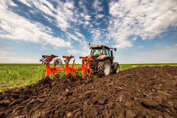 Red tractor is engaged in plowing the soil in a vast green farmland under a cloudy sky