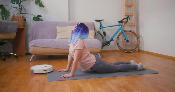 Young plus size latin hispanic teenager girl doing stretching when vacuum cleaner robot working around. Cozy home interior with indoor plants. Online fitness, urban jungle concept