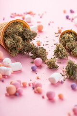 Obraz na płótnie Canvas Dry buds of medical marijuana lie on waffle ice cream cones on a pink background. There are candies and marshmello around. Alternative medical cannabis treatment