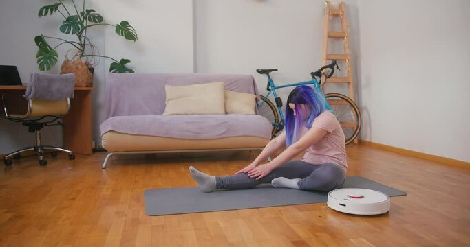 Young plus size latin hispanic teenager girl doing stretching when vacuum cleaner robot working around. Cozy home interior with indoor plants. Online fitness, urban jungle concept