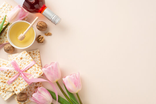 Top view picture of a Passover seder arrangement with matzah in a ribbon, a red wine bottle, walnuts, bowl of honey with a dipper spoon, and fresh pink tulips, on a pastel beige surface