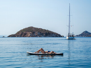 Beautiful blond woman in a bikini lying on a sup - stand up paddle sunbathing and enjoying serenity in the Mediterranean Sea. Sailing boat on the background. 