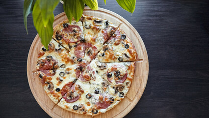 A freshly baked pepperoni and mushroom pizza with black olives and gooey melted cheese sits atop a circular wooden board against a dark table, partially framed by green leaves - Powered by Adobe