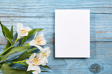 A portrait-oriented white blank card with a backdrop of blue painted wooden planks, adorned with blooming white Alstroemeria flowers