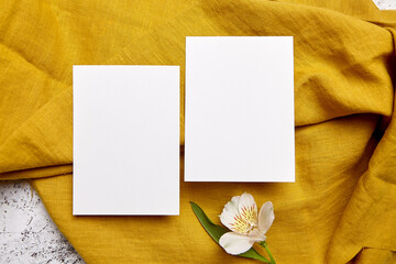 Two white blank greeting cards arranged on a mustard yellow textured linen with a single alstroemeria flower, top view, flat lay