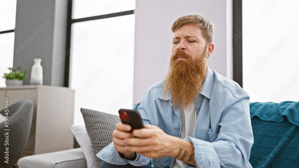 Wall mural focused young redhead man, chilling on his sofa indoors, engrossed in texting on his smartphone at h