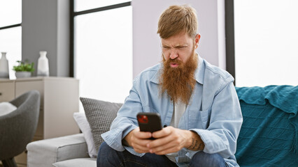 Focused young redhead man, chilling on his sofa indoors, engrossed in texting on his smartphone at his cozy apartment.