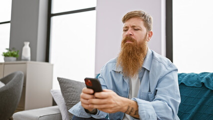 Focused young redhead man, chilling on his sofa indoors, engrossed in texting on his smartphone at...