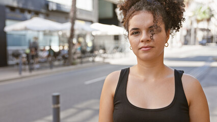 A young curly-haired hispanic woman stands confidently outdoors on a bustling urban street.