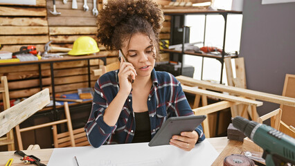 A young hispanic woman with curly hair is engaged in a phone conversation while holding a tablet in a woodwork workshop.