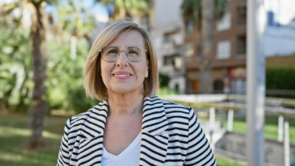 Smiling middle-aged woman with glasses posing outdoors in a city park with greenery.