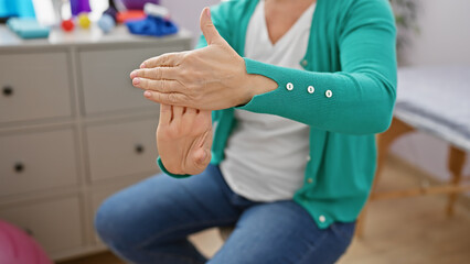 A middle-aged woman performs wrist strengthening exercises in a rehab clinic's interior.