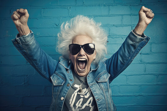 Exuberant senior lady with white hair and sunglasses, arms up, celebrating against a blue brick wall.
