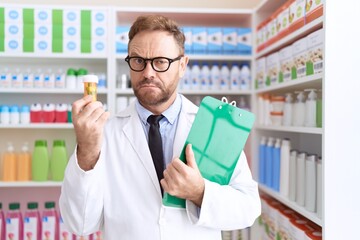 Middle age man with beard working at pharmacy drugstore holding pills clueless and confused...