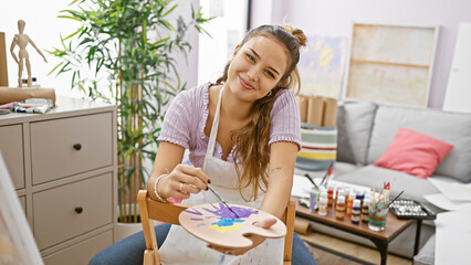 Smiling and confident, beautiful young hispanic woman artist skillfully holding her paintbrush and palette in bustling art studio setting