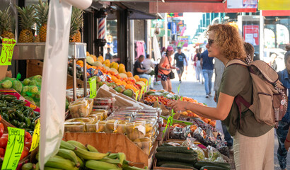 A woman examines fruits at a bustling street market, with vibrant fresh produce and prices...