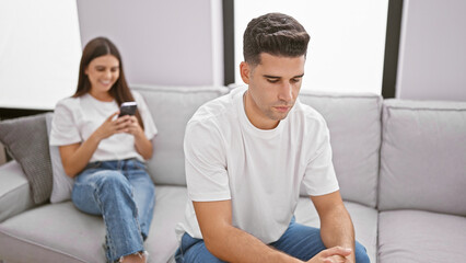 A young man and woman sitting apart on a sofa indoors, highlighting relationship tension with a blurred phone.