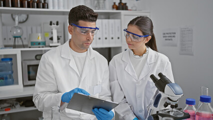 Man and woman in lab coats examine data on a tablet inside a science laboratory, depicting teamwork in research.