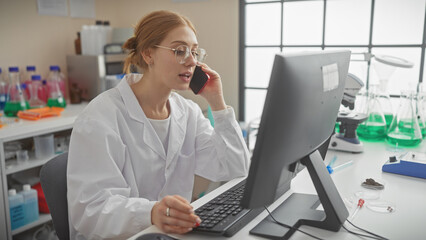 A professional woman scientist in a lab coat converses on the phone while working on a computer in...