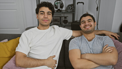 Two smiling men sitting closely on a grey sofa indoors, exuding a warm, friendly atmosphere.