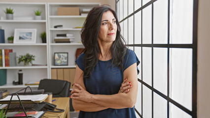 Confident hispanic mature woman with arms crossed standing in modern office, looking out window.