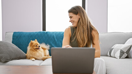Cheerful, young hispanic woman sitting on the sofa, using laptop with her playful dog, enjoying online technology in the comfort of her living room at home