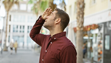 A young hispanic man with a beard looks upward, shielding his eyes, on a sunny outdoor city street.