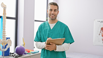 Portrait of a smiling young man in medical scrubs holding a clipboard in a clinic with anatomical...