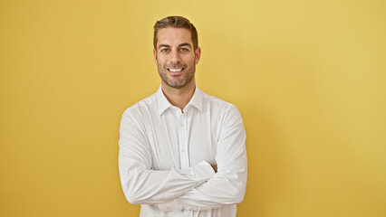 Confident young man with beard in white shirt posing with arms crossed against yellow background,...