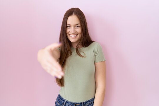 Beautiful brunette woman standing over pink background smiling friendly offering handshake as greeting and welcoming. successful business.