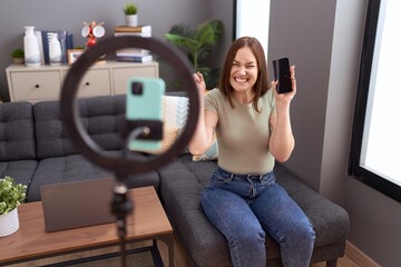 Beautiful brunette woman recording vlog tutorial with smartphone at home screaming proud,...