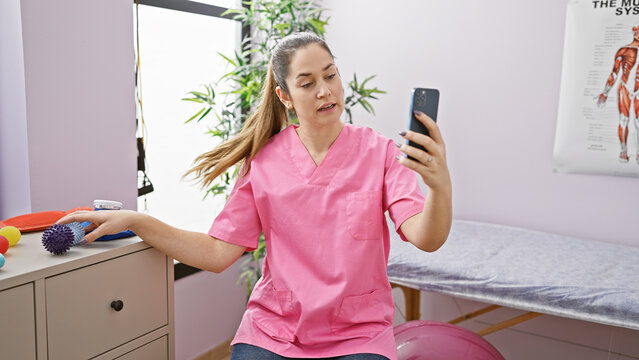 A woman in pink scrubs holds a phone for a video call in a therapy room with rehabilitation equipment.