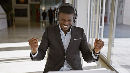 Handsome black man with headphones exults in a modern office setting, symbolizing success, motivation, and happiness.