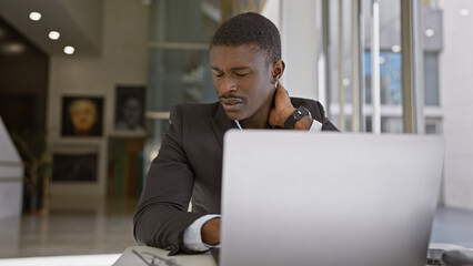 A professional african american man experiences neck pain while working on a laptop in a modern office setting.