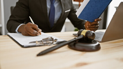 An african american man in a suit signs documents, reading a blue book beside a gavel in an office.