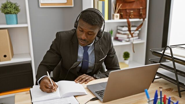 A focused african american man wearing a headset while writing notes in an office.