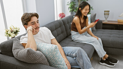 Disinterested man and smiling woman using a smartphone in a modern living room, depicting relationship dynamics.