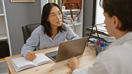 A woman and a man engage in a professional discussion with a laptop and notebook on a desk in a modern office setting.