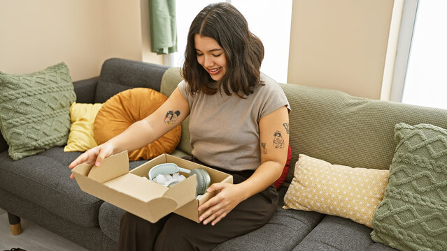 Smiling young hispanic woman unboxing headphones in a cozy living room setting, portraying leisure and technology at home.