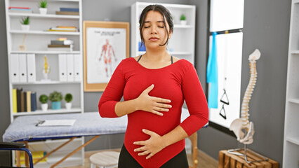 Young hispanic woman practices deep breathing techniques in a clinic's rehabilitation room,...
