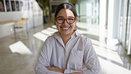 Smiling young hispanic woman professional posing confidently in a bright modern office interior.