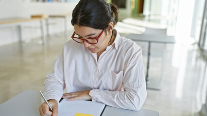 Focused hispanic woman writing on a notepad in a modern office setting, wearing glasses and a white shirt.